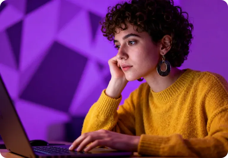 Curly-haired woman in a mustard sweater at a laptop, purple neon glow behind