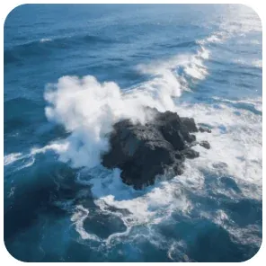 Aerial view of waves breaking white against dark rocks in deep blue sea